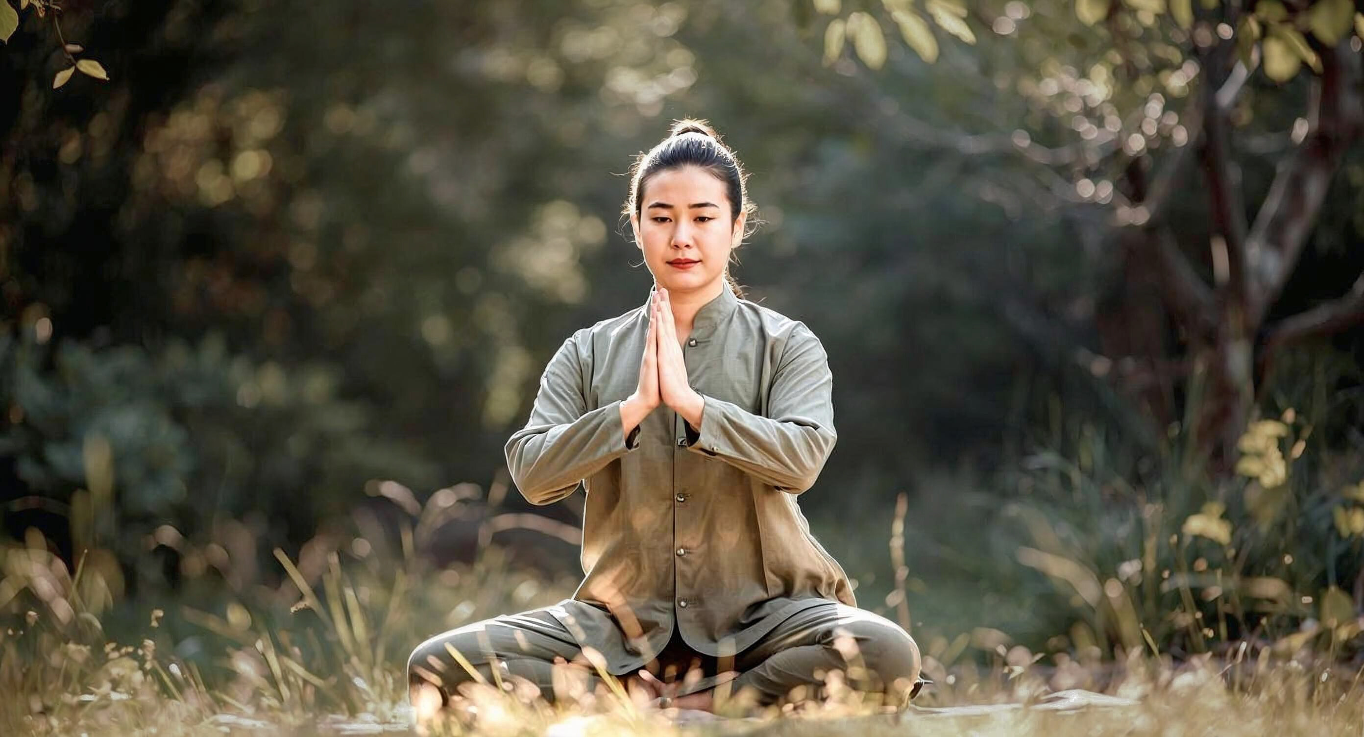 Eine asiatisch aussehende Frau sitzt im Schneidersitz in der Natur und meditiert.